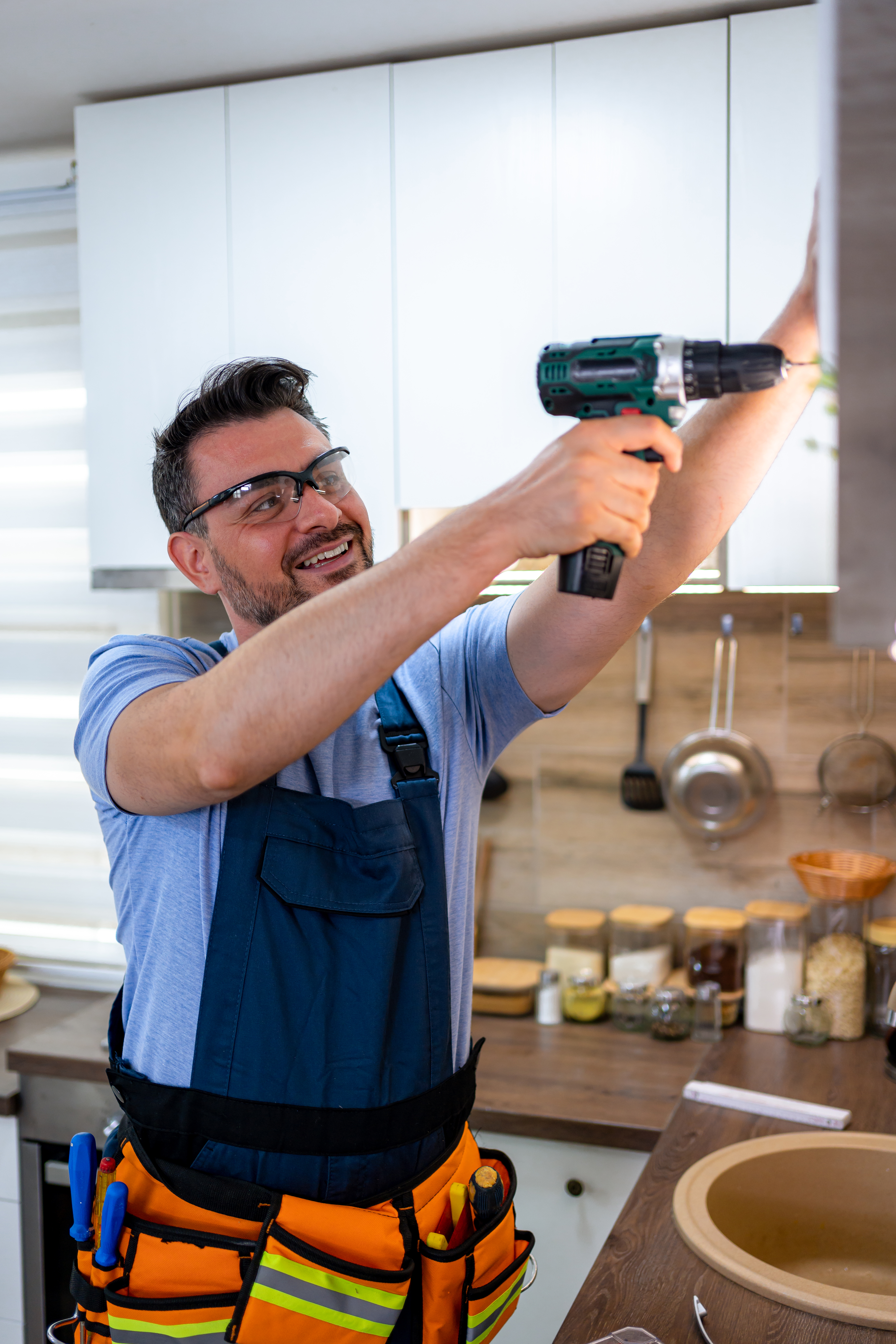A man smiles as he uses a power drill to install shelves in a bright kitchen.