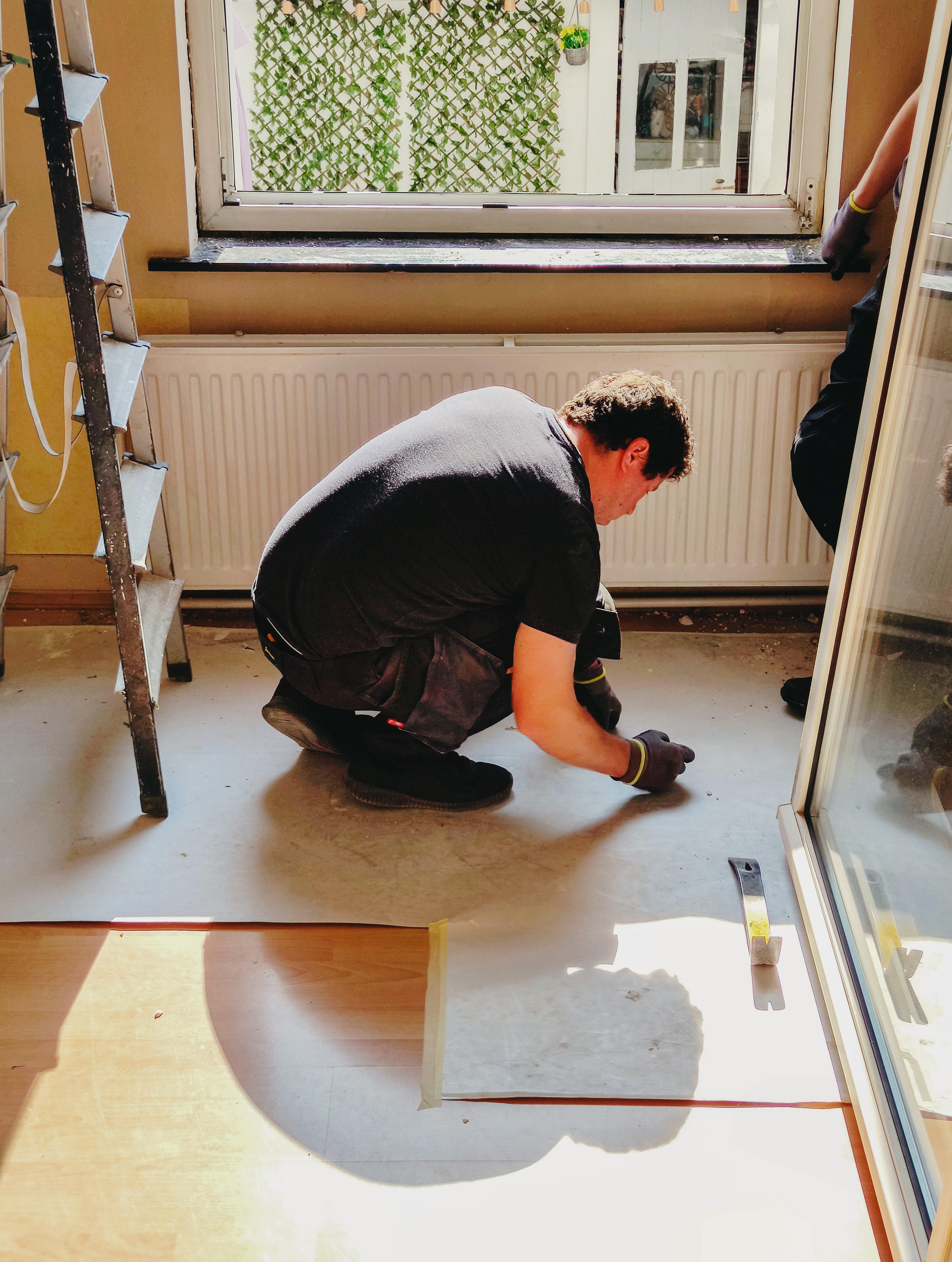 A mature Caucasian man in uniform squats and writes a note on a piece of paper on the floor before replacing an old window frame with a new one in a bedroom on a sunny spring day. Vertical side view, medium shot. Concept: repair and replacement of windows and window frames, using technology, mathematical calculations.